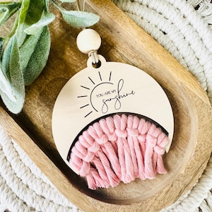 May include: A wooden sun-shaped ornament with a pink macrame fringe and the text "You are my sunshine" engraved on it.