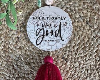 May include: Wooden car air freshener with a pink tassel. The round wooden disc has a floral design and the text "Hold Tightly to what is good - Romans 12:9"
