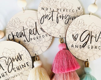 May include: Five wooden round ornaments with floral designs and inspirational quotes. Each ornament has a tassel attached. The quotes are "Not perfect just forgiven", "Created for a purpose", "Let your light shine Matthew 5:16", "Love and grace", and "Love and grace".