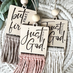 May include: Three wooden signs with tassels hanging from them. The signs have different religious quotes on them. The first sign says "but first pray." The second sign says "just trust God." The third sign says "trust God." The signs are all on a white background.