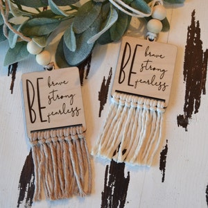 May include: Two wooden signs with the words "BE brave strong fearless" hanging from a string with wooden beads. Each sign has a fringe of natural colored yarn.