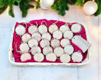 May include: A collection of wooden Christmas ornaments with various religious titles and phrases, displayed in a white basket on a pink cloth. Each ornament has a scalloped edge and a string for hanging. The ornaments are under a Christmas tree.