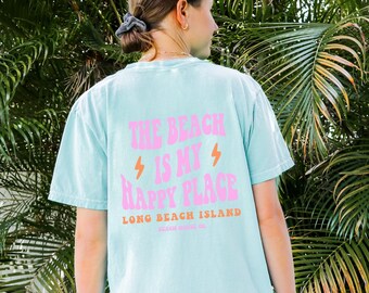 May include: A light blue t-shirt with a pink graphic that says "The Beach is my Happy Place" and "Long Beach Island" in orange text. The shirt is being worn by a person standing in front of a green leafy background.