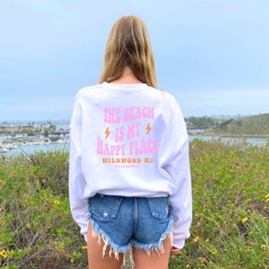 Puede incluir: Sudadera blanca de manga larga con texto rosa que dice "The Beach is my Happy Place" y "Wildwood NJ". La sudadera la lleva puesta una persona de pie frente a una masa de agua.