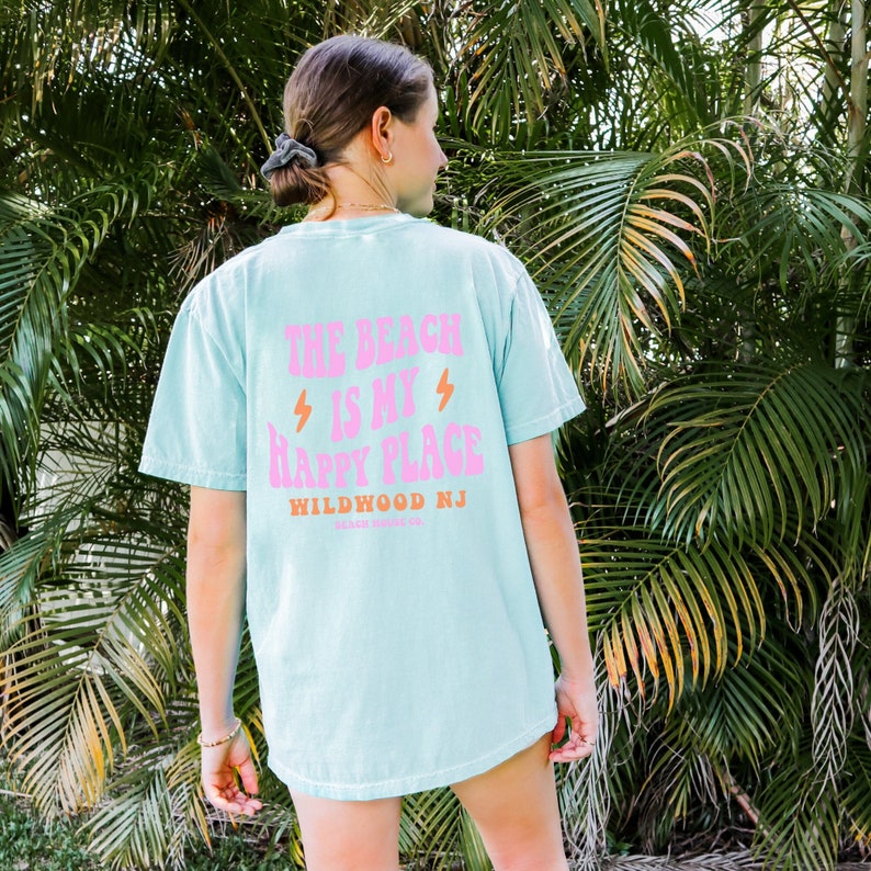 K&ouml;nnte beinhalten: Ein hellblaues T-Shirt mit einer pinkfarbenen Grafik, auf der "The Beach is my Happy Place" und "Wildwood NJ" in Orange steht. Das Shirt wird von einer Person getragen, die vor einem gr&uuml;nen, belaubten Hintergrund steht.