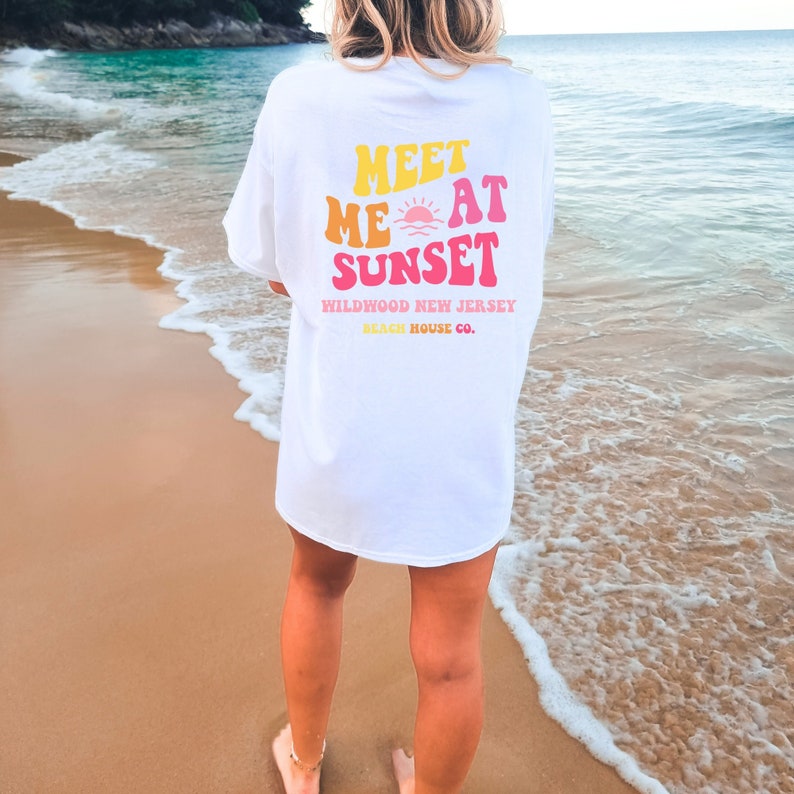 Puede incluir: Camiseta blanca con un gr&aacute;fico que dice "Meet Me At Sunset" en una fuente retro con un gr&aacute;fico de puesta de sol. El texto debajo del gr&aacute;fico dice "Wildwood New Jersey Beach House Co."