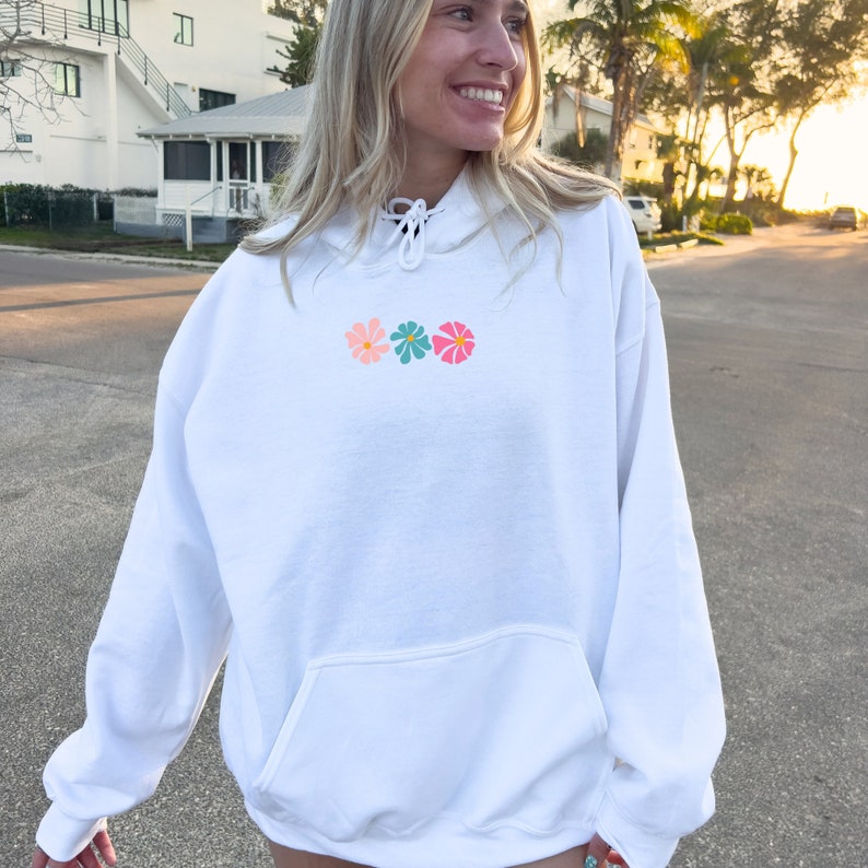 May include: White hoodie with a drawstring hood and a pocket. The hoodie has a small embroidered design of three colorful flowers.