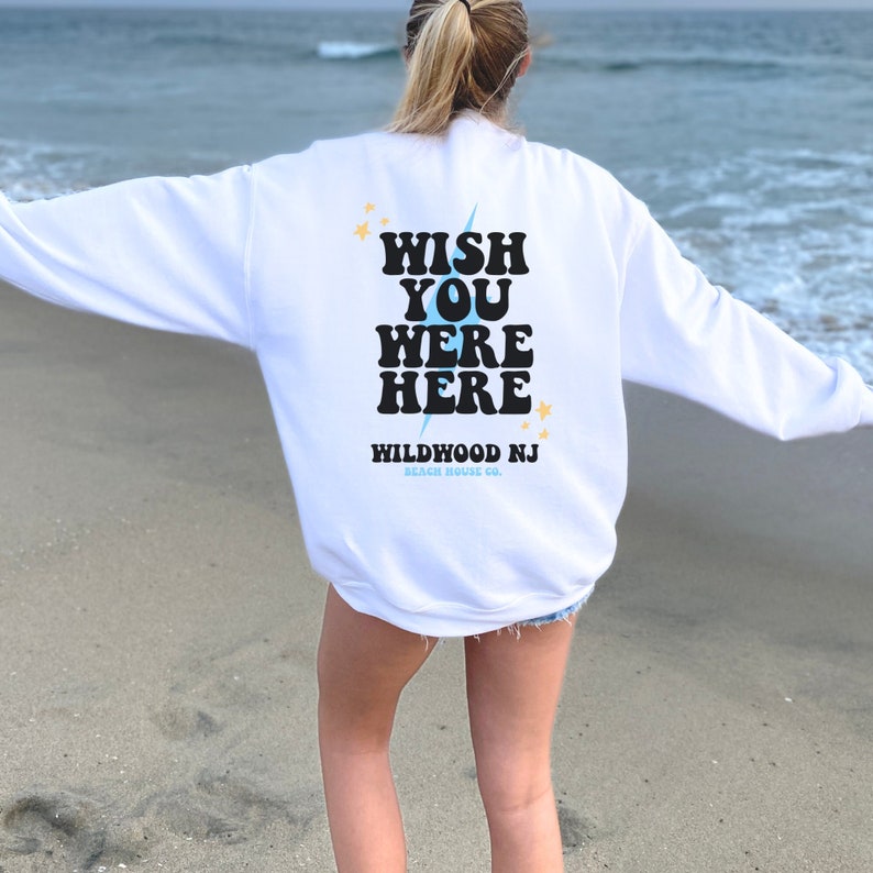 May include: White sweatshirt with the text "WISH YOU WERE HERE" in black retro font with a blue lightning bolt and yellow stars. Below the text is "WILDWOOD NJ BEACH HOUSE CO."