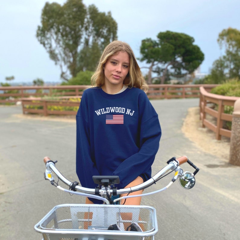 Puede incluir: Una persona joven lleva una sudadera azul marino con el texto "WILDWOOD NJ" y un peque&ntilde;o gr&aacute;fico de la bandera estadounidense. Est&aacute; montando una bicicleta plateada con una cesta.