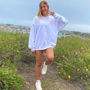 Puede incluir: Una sudadera blanca con un gr&aacute;fico de rayo rosa y la palabra "⚡️⚡️⚡️" impresa en la parte delantera. La sudadera la lleva puesta una persona de pie en un campo de flores amarillas.