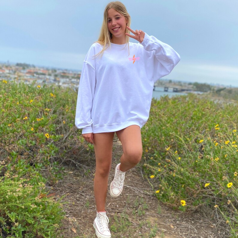 Puede incluir: Una sudadera blanca con un gr&aacute;fico de rayo rosa y la palabra "⚡️⚡️⚡️" impresa en la parte delantera. La sudadera la lleva puesta una persona de pie en un campo de flores amarillas.
