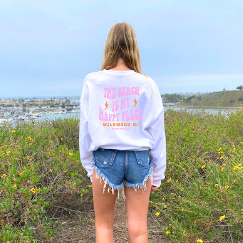 Puede incluir: Sudadera blanca de manga larga con texto rosa que dice "The Beach is my Happy Place" y "Wildwood NJ". La sudadera la lleva puesta una persona de pie frente a una masa de agua.