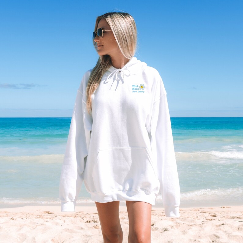 K&ouml;nnte beinhalten: Wei&szlig;er Kapuzenpullover mit einem blauen und gelben Blumenmotiv und dem Text "Wild Blood New Jersey". Das Model tr&auml;gt den Pullover am Strand.