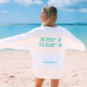 K&ouml;nnte beinhalten: Wei&szlig;er Pullover mit den Koordinaten "38.9918&deg; N 74.8149&deg; W" und dem Text "Beach House Co." auf der R&uuml;ckseite.