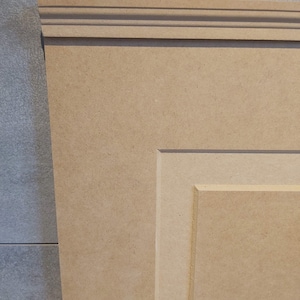 May include: A close-up of a light brown wooden panel with a raised, decorative frame. The panel has a smooth, unfinished surface and is made of medium-density fiberboard (MDF).