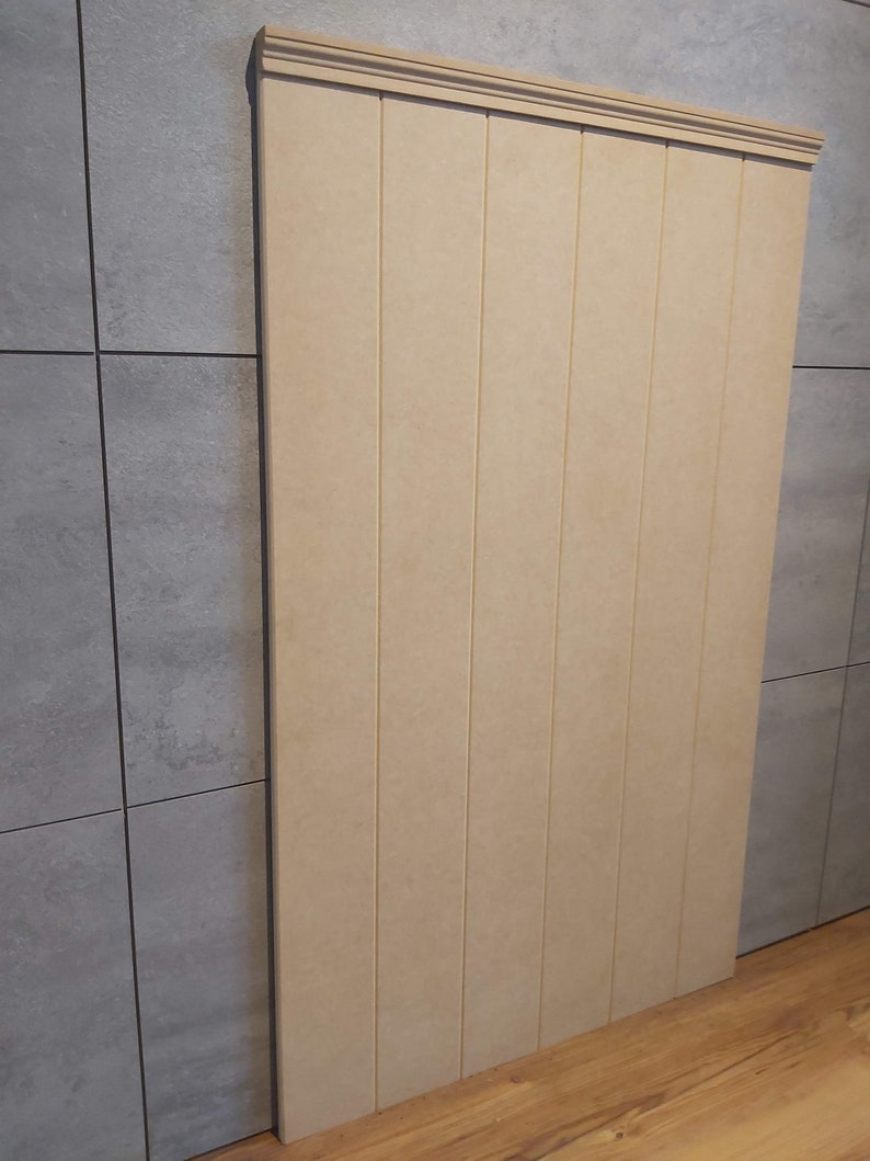 May include: A vertical, rectangular wooden panel with a decorative top molding. The panel is composed of several vertical planks, each with a narrow groove. The wood has a light beige color, and the panel is positioned against a gray tiled wall.