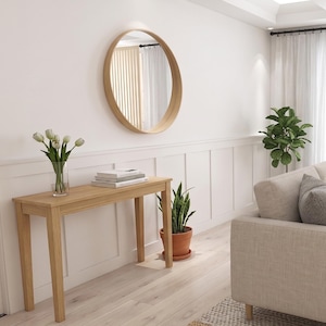Peut inclure: Un miroir mural rond avec un cadre en bois clair est suspendu au-dessus d'une console rectangulaire en bois. La table supporte un vase en verre transparent avec des tulipes blanches et une pile de livres. Une plante serpent est pos&eacute;e au sol.