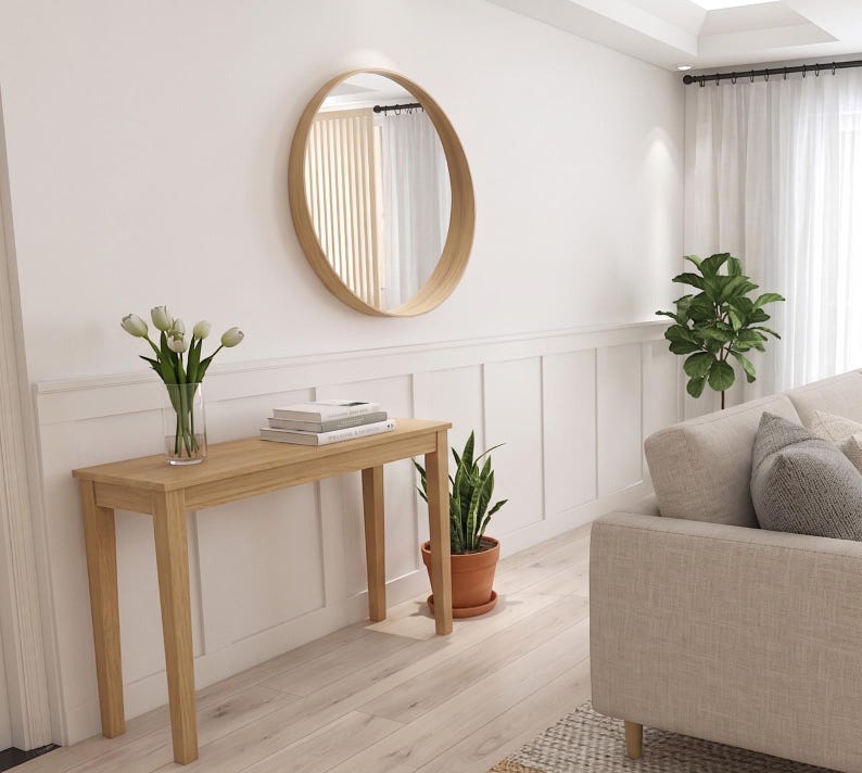 Peut inclure: Un miroir mural rond avec un cadre en bois clair est suspendu au-dessus d'une console rectangulaire en bois. La table supporte un vase en verre transparent avec des tulipes blanches et une pile de livres. Une plante serpent est pos&eacute;e au sol.