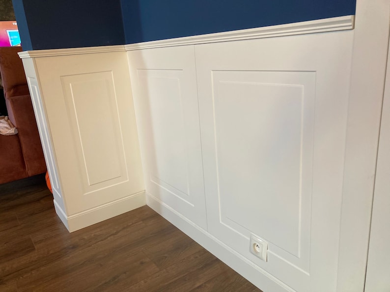 May include: White wainscoting with a raised panel design, painted in a glossy white finish. The wainscoting is installed on a wall with a dark blue accent color above. An electrical outlet is visible on the lower panel.