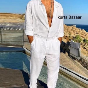 Op de afbeelding: Een witte linnen jumpsuit met lange mouwen en een knoopsluiting aan de voorkant. De jumpsuit heeft een ontspannen pasvorm en een licht ingekort been. Het model draagt de jumpsuit met een zilveren ketting. De tekst "kurta Bazaar" is zichtbaar in de rechterbovenhoek van de afbeelding.