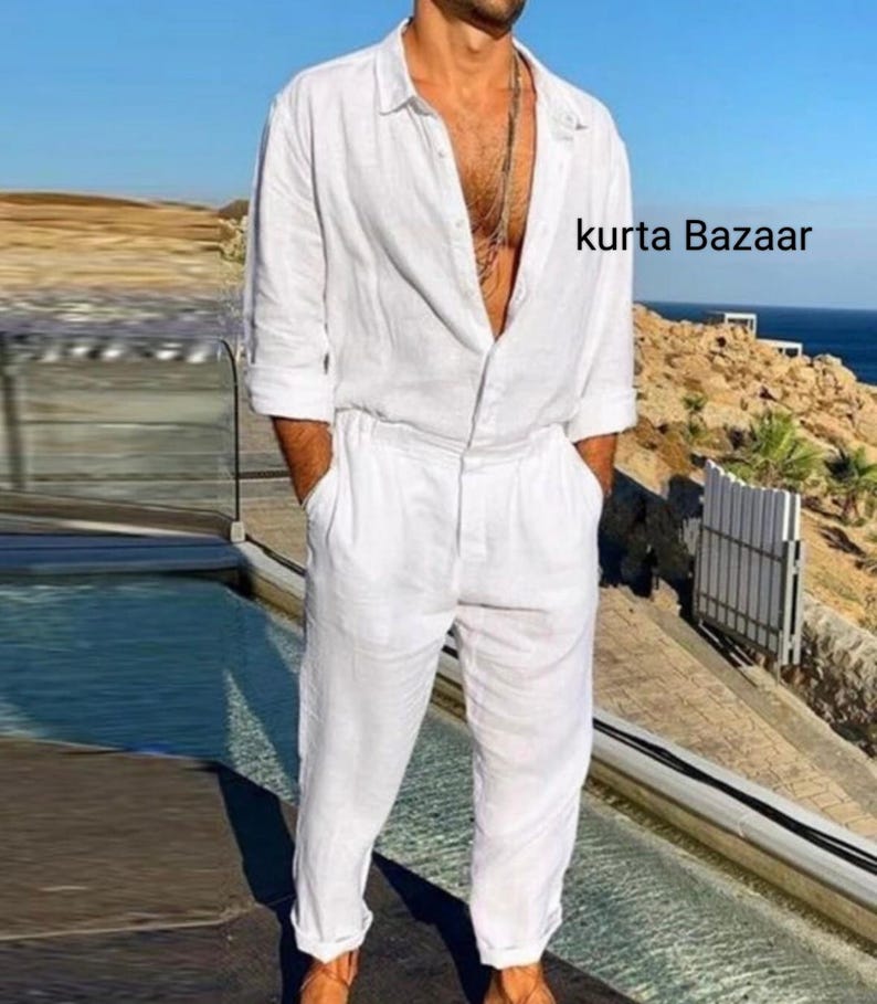 Op de afbeelding: Een witte linnen jumpsuit met lange mouwen en een knoopsluiting aan de voorkant. De jumpsuit heeft een ontspannen pasvorm en een licht ingekort been. Het model draagt de jumpsuit met een zilveren ketting. De tekst "kurta Bazaar" is zichtbaar in de rechterbovenhoek van de afbeelding.
