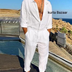 Op de afbeelding: Een witte linnen jumpsuit met een lang mouw shirt en een knoopsluiting aan de voorkant. De jumpsuit heeft een ontspannen pasvorm en een licht ingekort been. Het model draagt de jumpsuit met een zilveren ketting en sandalen. De tekst "kurta Bazaar" is zichtbaar in de afbeelding.
