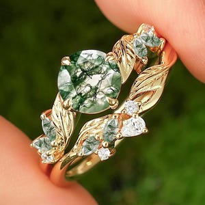 May include: A gold ring set featuring a round, green moss agate stone and a leaf-inspired band. The set includes a matching band with smaller green stones and clear accent stones. The rings are held against a blurred green background.
