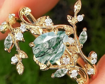 May include: A gold ring featuring a large, square-cut, moss agate gemstone with green inclusions. The ring is adorned with delicate gold leaves and small, sparkling white gemstones, creating a nature-inspired design.