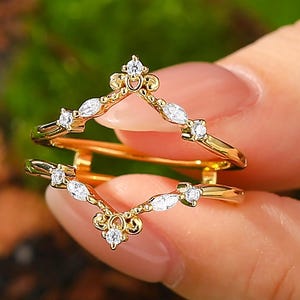 May include: A gold-colored ring guard with a V-shaped design, adorned with small, clear gemstones. The ring guard features a delicate, ornate pattern and is held against a blurred green background.