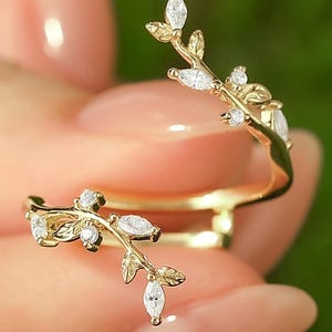 Könnte beinhalten: Ein goldfarbener Ring mit Blattdesign. Der Ring hat kleine, klare Edelsteine, die entlang der Zweige der Blätter gesetzt sind. Der Ring umschließt den Finger und erzeugt einen einzigartigen und eleganten Look.