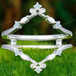 May include: A silver ring guard featuring two chevron-shaped bands connected by a bridge. The design includes small, round, light blue stones and marquise-cut white opals set along the V-shaped metal frame. The text MAYDAYJEWELRY is visible.