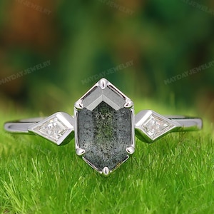 May include: A silver-toned ring featuring a central hexagonal-cut dark gray stone with speckled inclusions, flanked by two small kite-shaped clear stones on a polished metal band. The text MAYDAYJEWELRY is visible in the background.