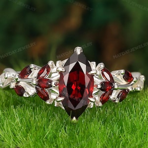 May include: A silver ring featuring a central, marquise-cut, deep red gemstone. The band is embellished with smaller red gemstones and silver leaf detailing. The ring is displayed on green grass. The text "MAYDAYJEWELRY" is visible.
