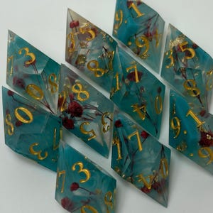 May include: A collection of ten teal resin dice, each with gold numerals and dried flower inclusions. These polyhedral dice are designed for role-playing games. The dice are various shapes and sizes, offering a unique aesthetic for tabletop gaming.