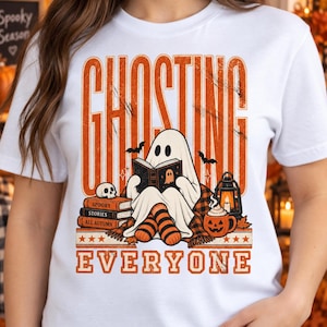 Könnte beinhalten: Ein weißes T-Shirt mit einer Grafik eines geisterhaften Wesens, das ein Buch liest, und dem Text GHOSTING EVERYONE. Das Design zeigt einen Stapel Bücher mit der Aufschrift Spooky Stories All Autumn, einen Totenkopf, eine Laterne, eine Kürbistasse und orange gestreifte Socken.
