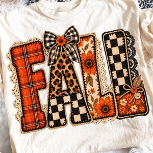 May include: A cream-colored t-shirt featuring the word FALL in large block letters. Each letter has a different pattern: red plaid, leopard print, black and white checkerboard, and sunflower print. A black and white plaid bow sits atop the A.