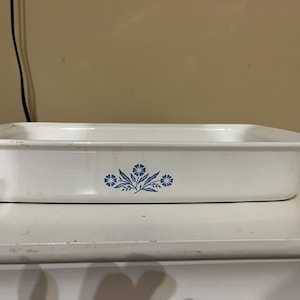 以下が含まれることがあります： 青い花の模様が入った白い長方形のベーキングディッシュ。 陶器製で、少し使い古した外観です。 花柄はディッシュの前面中央に配置されています。
