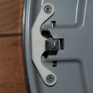 May include: A close-up of a white plastic latch with two screws. The latch is attached to a gray metal surface.