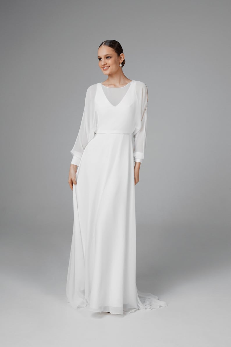 May include: A white wedding dress with a flowing skirt and sheer, long sleeves. The dress features a V-neckline and a fitted bodice. The sleeves are slightly flared at the wrists. The dress is made of a soft, lightweight fabric.