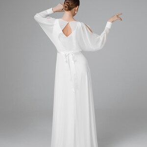 May include: A white, flowing gown with long, sheer sleeves and a keyhole back. The dress features a cinched waist with a long, matching sash. The model's hair is styled in a bun, and she wears pearl earrings. The dress is suitable for a wedding or formal event.