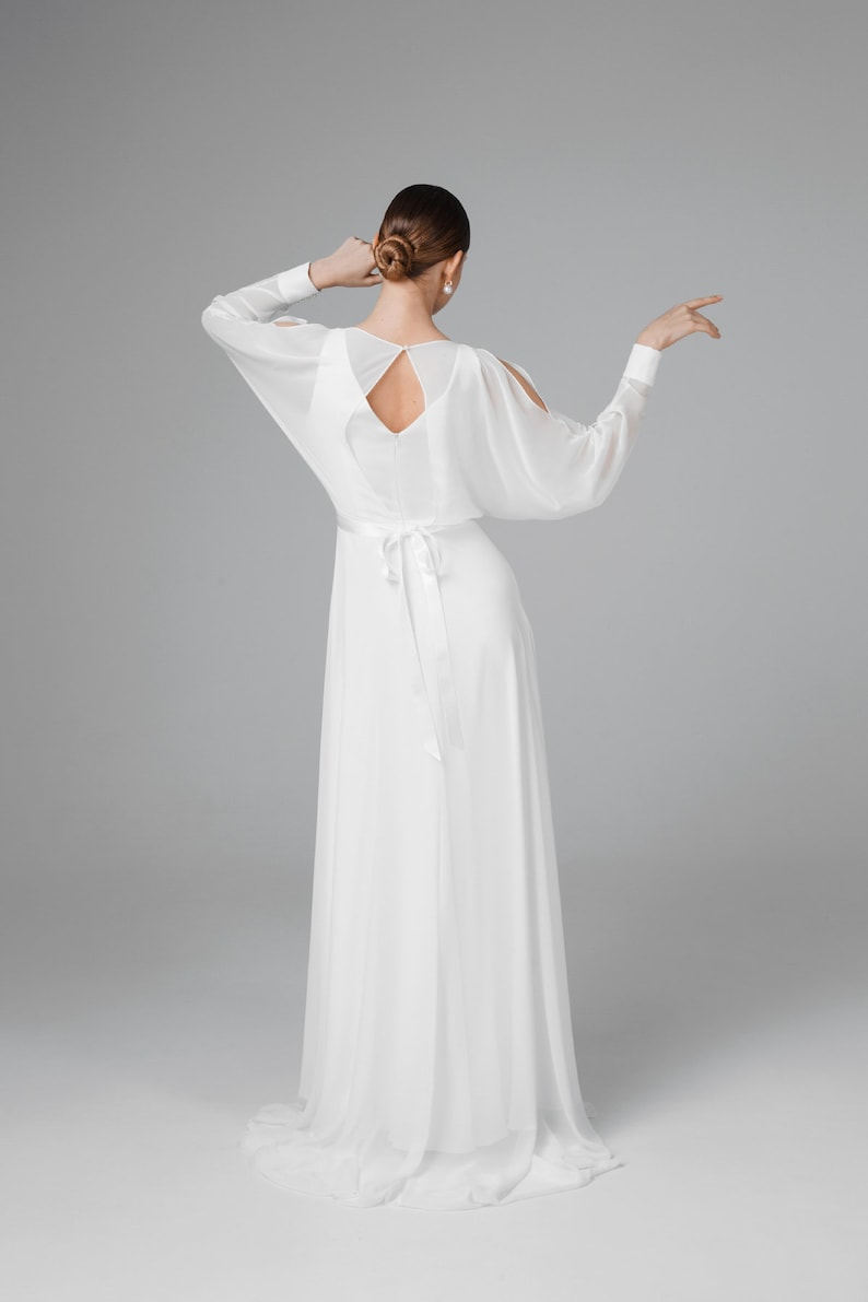 May include: A white, flowing gown with long, sheer sleeves and a keyhole back. The dress features a cinched waist with a long, matching sash. The model's hair is styled in a bun, and she wears pearl earrings. The dress is suitable for a wedding or formal event.