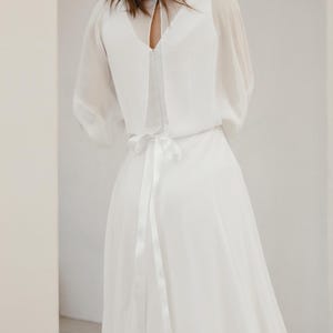 May include: A white wedding dress with a flowing skirt and sheer sleeves. The dress features a keyhole back and a ribbon tie at the waist. The model is facing away from the camera.