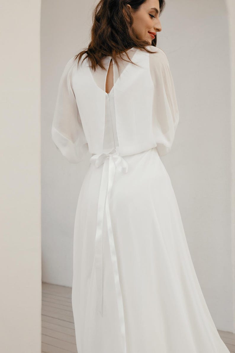May include: A white wedding dress with a flowing skirt and sheer sleeves. The dress features a keyhole back and a ribbon tie at the waist. The model is facing away from the camera.