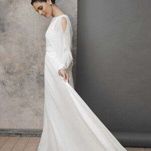 May include: A white wedding dress with long sleeves and a flowing skirt. The dress features a boat neckline and cut-out details on the upper arms. The bride is holding the skirt of the dress, which has a long train.