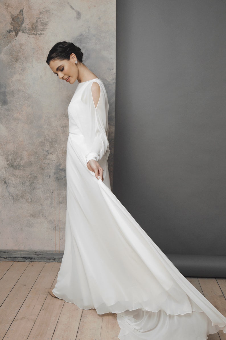 May include: A white wedding dress with long sleeves and a flowing skirt. The dress features a boat neckline and cut-out details on the upper arms. The bride is holding the skirt of the dress, which has a long train.