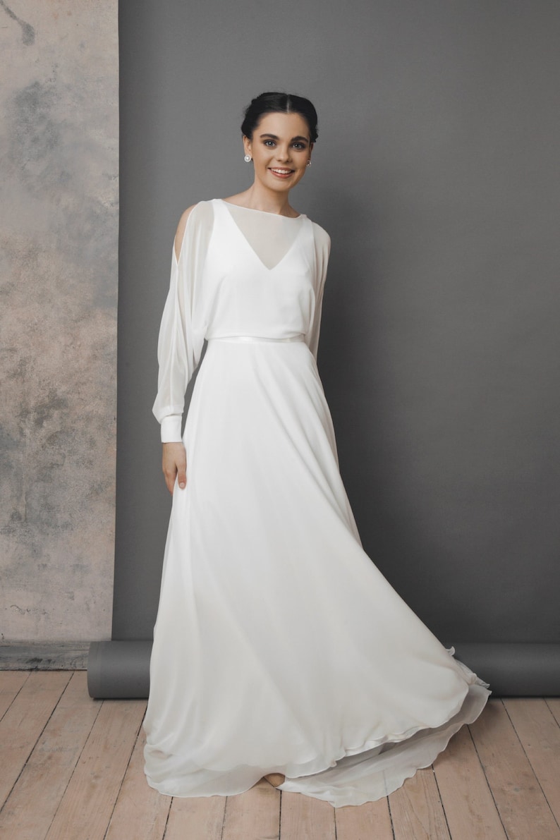 May include: A white wedding dress with a flowing skirt and sheer sleeves. The dress features a V-neckline and a delicate belt at the waist. The model is smiling, and the dress appears to be made of a lightweight fabric.