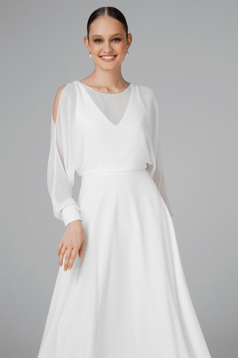 May include: A white wedding dress with a flowing skirt and sheer, long sleeves. The dress features a V-neckline and cut-out shoulder details. The model is smiling and wearing pearl earrings.