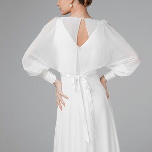 May include: A white wedding dress with a flowing skirt and long sleeves. The dress features a keyhole back and a satin tie at the waist. The sleeves are sheer and have button details at the cuffs. The model's hair is styled in a bun.