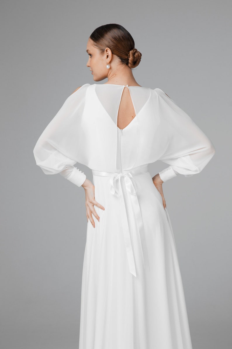 May include: A white wedding dress with a flowing skirt and long sleeves. The dress features a keyhole back and a satin tie at the waist. The sleeves are sheer and have button details at the cuffs. The model's hair is styled in a bun.