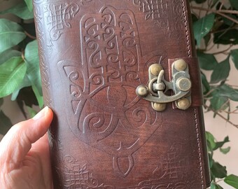 May include: A brown leather journal with a hamsa hand design embossed on the front cover. The journal has a brass clasp closure and is decorated with intricate patterns.
